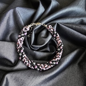 May include: A beaded bracelet with a silver clasp. The bracelet features small, round beads in black, pink, and grey. The bracelet is displayed on a dark grey, silky fabric.