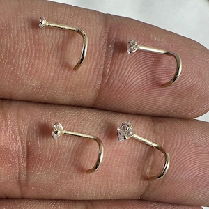 May include: Four gold nose rings with clear gemstones. Two rings have round gemstones and two rings have square gemstones.