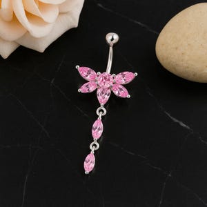 Puede incluir: Un piercing para el ombligo plateado con un diseño floral rosa y una cadena de gemas rosas. La flor tiene seis pétalos alrededor de una gema central. Las gemas son ovaladas y están engastadas en metal plateado.