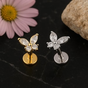 Op de afbeelding: Twee vlindervormige oorstekers. Eén is goudkleurig, de andere zilverkleurig. Elk heeft vier heldere, sprankelende edelstenen. De oorbellen worden gepresenteerd op een zwart oppervlak, met een roze bloem en een steen op de achtergrond.