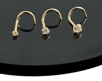 Puede incluir: Tres anillos de nariz de oro con pequeñas piedras preciosas transparentes. Los anillos están sobre una superficie negra.