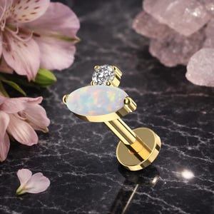 May include: A gold-coloured labret stud with a white opal marquise-shaped stone and a small, clear, round gemstone. The stud is set against a dark, marbled surface, with pink flowers in the background.