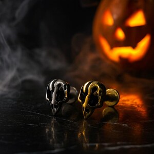 May include: Two skull-shaped cufflinks, one silver and one gold, are displayed on a dark surface. The background features a lit jack-o'-lantern and wisps of smoke, creating a Halloween theme. The cufflinks are detailed and have a gothic aesthetic.