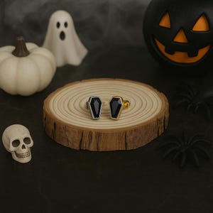 Puede incluir: Dos anillos con forma de ataúd con piedras negras, uno con una banda plateada y otro con una banda dorada, presentados sobre una rodaja de madera. Decoraciones de Halloween: calabaza, fantasma, calavera y arañas.