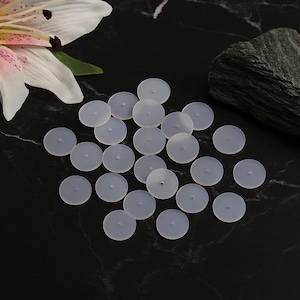 Op de afbeelding: Een verzameling kleine, doorschijnende, ronde siliconen oorbelruggen verspreid over een zwart oppervlak. De oorbelruggen hebben een klein gaatje in het midden. Een witte en roze leliebloem bevindt zich in de linkerbovenhoek.