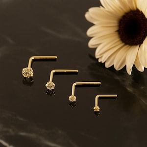 May include: Four gold-colored nose studs with different sized round gemstones. The studs are arranged in a row on a dark, reflective surface. A white and yellow flower is in the background.