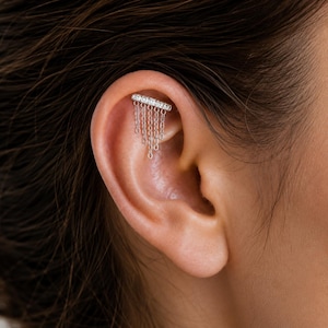 Può includere: Un orecchino da cartilagine color argento con una barra orizzontale incastonata con piccole pietre chiare. Cinque delicate catenelle argentate di lunghezze variabili pendono dalla barra, creando un effetto a cascata nella cartilagine.