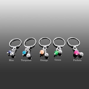 Puede incluir: Cinco anillos de plata con colgantes. Cada colgante presenta una cuenta de color y una pequeña campana plateada. Los colores incluyen azul, turquesa, naranja, verde y fucsia. Las palabras de cada color están escritas debajo.