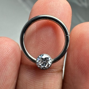 May include: A silver circular body piercing with a clear gem in the center.