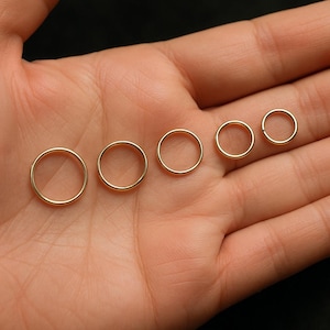 May include: Five gold-colored hoop earrings of varying sizes are displayed on a person's palm. The earrings are circular and have a smooth, polished finish. The smallest earring is about 0.25 inches in diameter, while the largest is about 0.75 inches.