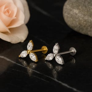 May include: Two pieces of jewelry, each featuring a three-petal design with clear, marquise-cut stones. One is gold-toned, the other silver-toned, both with a flat back for piercing. The jewelry is displayed on a black surface.