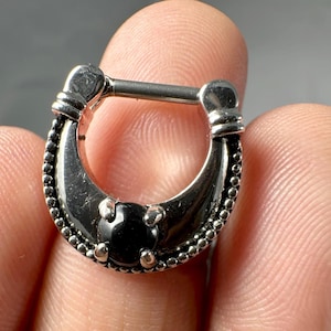 Puede incluir: Un piercing para el cuerpo de plata en forma de herradura con una piedra preciosa negra en el centro. El piercing tiene un borde con cuentas y una abertura con bisagra.