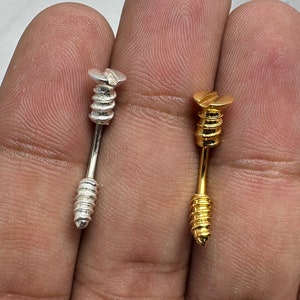 May include: Two screw-shaped body jewellery pieces, one silver and one gold, are shown on a hand. The jewellery is designed to look like a screw.