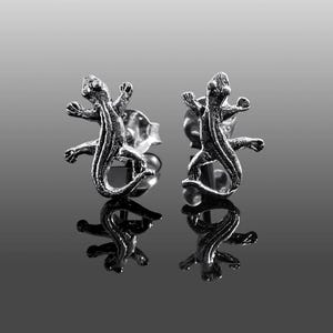 May include: A pair of silver-toned stud earrings, each shaped like a lizard with outstretched limbs. The earrings are highly detailed, with visible scales and a reflective surface. The lizard earrings are displayed on a reflective surface.