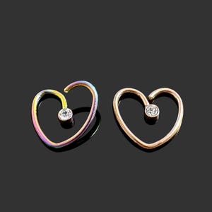 Puede incluir: Dos pendientes de aro en forma de corazón con una piedra preciosa transparente en el centro. Un pendiente es de color arcoíris y el otro es de oro rosa.
