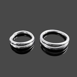 以下が含まれることがあります： シルバーカラーのヒンジ付きフープピアス2つ。各イヤリングは、滑らかで反射性の表面を持つトリプルバンドデザインです。イヤリングは濃い灰色の背景に表示され、金属的な輝きを際立たせています。
