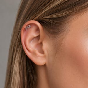 May include: A silver-toned ear cartilage stud featuring three small, clear, rectangular-cut stones arranged in a row. The jewelry is positioned in the upper cartilage area of the ear, showcasing a metallic setting with a secure post backing.