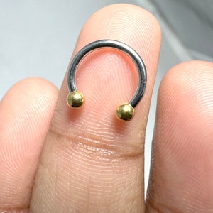 May include: A silver horseshoe-shaped ring with gold-colored balls on each end. The ring is made of metal and is a type of body jewelry.