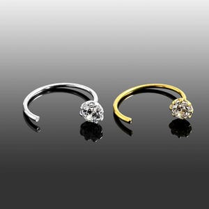 Puede incluir: Dos piercings de nariz, uno plateado y otro dorado, cada uno con una pequeña y brillante gema redonda. Los anillos tienen un diseño curvo y abierto. El fondo es un degradado de negro y gris.