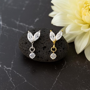 Könnte beinhalten: Zwei blattförmige Ohrringe mit klaren, funkelnden Edelsteinen. Ein Ohrring ist silberfarben, der andere goldfarben. Jeder hat einen hängenden, quadratisch geschliffenen Edelstein. Die Ohrringe liegen auf einem dunklen, strukturierten Stein.