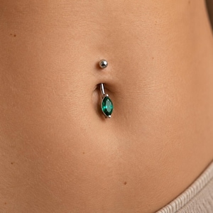 Könnte beinhalten: Ein silberfarbener Bauchnabelpiercing-Ring mit einem smaragdgrünen Edelstein im Marquise-Schliff. Der Schmuck besteht aus einem gebogenen Stab mit einer kleinen polierten Metallkugel oben und einem größeren, facettierten grünen Stein unten.