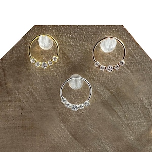 Peut inclure: Trois boucles d'oreilles créoles avec des pierres précieuses transparentes. L'une est dorée, l'autre argentée et la dernière dorée. Chaque boucle d'oreille comporte une rangée de cinq pierres précieuses. Les boucles d'oreilles sont exposées sur une surface en bois.
