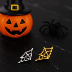 Może przedstawiać: Obraz o tematyce Halloween z małą pomarańczową dynią z czarnym kapeluszem czarownicy i wyrzeźbioną twarzą. Dwie ozdoby w kształcie pajęczyny, jedna srebrna i jedna złota, znajdują się na pierwszym planie.