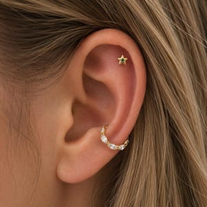 May include: A gold star-shaped stud earring with a small green center stone positioned in the upper ear cartilage, paired with a gold hoop earring featuring a row of clear marquise-cut stones worn in the lower ear lobe.
