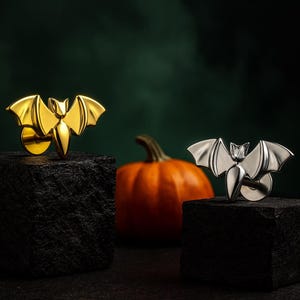 Könnte beinhalten: Zwei fledermausförmige Anstecknadeln, eine goldene und eine silberne, werden auf schwarzen Steinblöcken präsentiert. Im Hintergrund befindet sich ein orangefarbener Kürbis vor einem dunkelgrünen Hintergrund. Halloween-Accessoires.