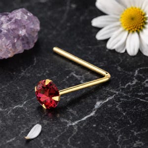 Puede incluir: Un piercing de nariz dorado con una gema roja vibrante. La joya en forma de L está colocada sobre una superficie de mármol negro, con una margarita y un cristal de amatista en el fondo. La gema es de forma ovalada.