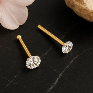Peut inclure: Deux clous de nez dorés avec des pierres de zircone cubique claires de coupe ovale. Les clous ont une tige droite et une petite boule à l'extrémité opposée de la pierre. Le fond est une surface sombre et texturée.