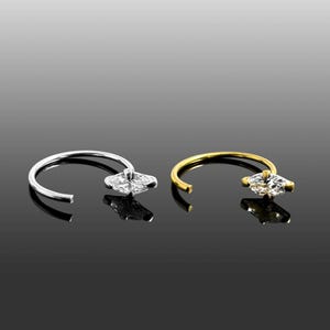Puede incluir: Se muestran dos piercings nasales, uno plateado y otro dorado. El anillo plateado presenta una gema en forma de flor, mientras que el anillo dorado tiene una gema con corte marquesa. Ambos anillos tienen un diseño curvo.