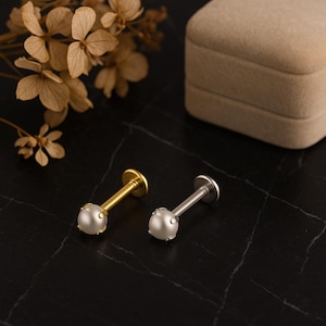 May include: Two lip or cartilage earrings with pearl-like studs. One earring is gold-colored, the other is silver-colored. Both have a flat back and a post. The earrings are displayed on a black, textured surface.