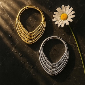 Puede incluir: Dos colgantes ovalados de metal, uno dorado y otro plateado, con líneas curvas superpuestas, se muestran sobre una superficie oscura y texturizada. También hay una margarita blanca con centro amarillo. Joyas.