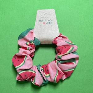 Peut inclure: Un chouchou en tissu rose avec un imprimé pastèque. Le chouchou a une étiquette qui dit "Fait main avec amour".