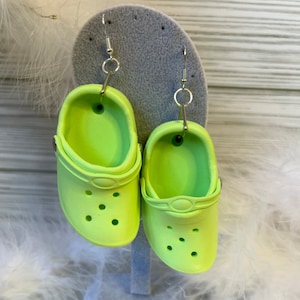 May include: A pair of neon green clog earrings. Each earring is a miniature replica of the popular shoe style, complete with the signature holes and a silver hook for the ear. The earrings are displayed on a gray earring stand.