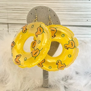 May include: A pair of novelty earrings shaped like yellow inflatable rings, adorned with cartoon duck illustrations. The earrings have gold-colored hooks and are displayed on a gray stand, evoking a playful, summery theme.