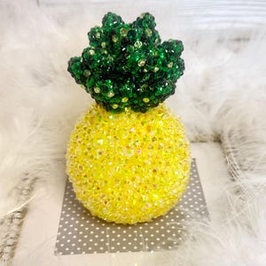 May include: A decorative pineapple figurine covered in yellow and green sparkling crystals. The pineapple has a yellow body and a green crown. The figurine is placed on a gray and white polka dot surface.