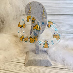 May include: Clear, round earrings with a yellow duck design. The earrings are shaped like inflatable rings, with gold-colored hooks. The ducks are painted with orange beaks and blue accents. The earrings are displayed on a gray stand.