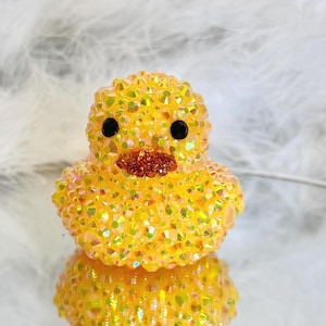 May include: A yellow, bedazzled duck figurine with black eyes and a reddish-brown beak. The duck is covered in iridescent crystals, creating a sparkling effect. The figurine is reflected in a shiny surface.
