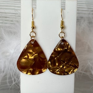 May include: A pair of gold-colored teardrop earrings. The earrings have a textured surface and are suspended from gold-colored ear wires. The earrings are displayed on a white stand against a soft, white background.