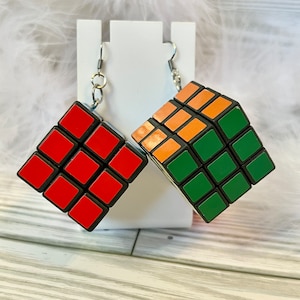 May include: A pair of Rubik's Cube earrings. One earring features red squares, the other has orange and green squares. The earrings are suspended from silver-colored fish hook ear wires.