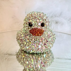 May include: A bedazzled duck figurine. The duck is covered in iridescent rhinestones, with black eyes and an orange beak. The figurine is reflected on a mirrored surface, with white feathers in the background.