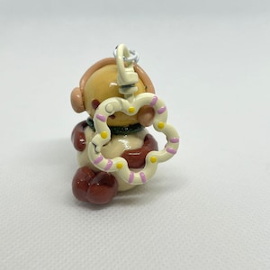 May include: A small, cream-coloured bear figurine with brown accents, wearing headphones and a green scarf. It holds a heart-shaped charm with pink and yellow polka dots. The charm is attached to a silver-coloured clasp, suggesting a keychain or accessory.