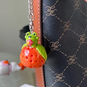 May include: A novelty keychain with a green frog sitting on a red strawberry. The frog has yellow eyes and a pink flower. The keychain is attached to a silver chain. The background includes a patterned handbag.