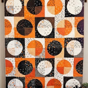 May include: A rectangular quilt featuring a repeating pattern of large circles composed of four quadrants. The fabric displays a Halloween theme with orange, black, and white colors, incorporating prints of bats, spiderwebs, and text.