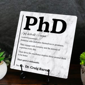 May include: A square white sign with a grey marble pattern featuring the black text PhD, its pronunciation, and a definition describing someone who dedicates themselves to greatness, with the name Dr. Craig Barn at the bottom.