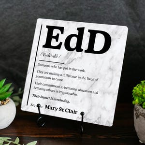 May include: A square white plaque with a grey marble pattern featuring the black text EdD, a phonetic pronunciation guide, and a definition describing someone who has put in the work to make a difference in education, ending with See also: Mary St Clair.