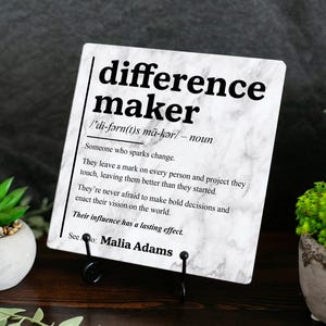 May include: A square white marble-patterned plaque featuring black text that defines the term difference maker as someone who sparks change, leaves a mark on others, and enacts a vision, with the name Malia Adams printed at the bottom.