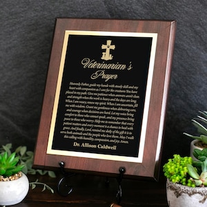 May include: A rectangular wooden plaque with a dark brown finish and a gold-bordered black center plate. The plate features a cross with a paw print, the title Veterinarian's Prayer, the full prayer text, and the name Dr. Allison Caldwell.